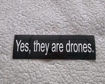 May include: Black and white sticker with the text "Yes, they are drones."