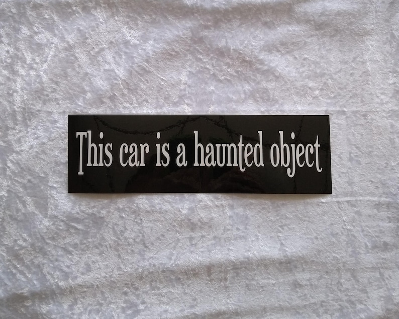 Rectangular sticker with black background and white lettering. The text reads this car is a haunted object.