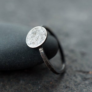 Op de afbeelding: Een zilveren ring met een ronde, getextureerde, zilveren bovenkant. De ring ligt op een donkergrijze steen.
