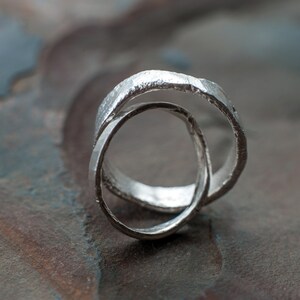 May include: Two silver rings intertwined, creating a unique and modern design. The rings are textured and have a slightly rough finish.