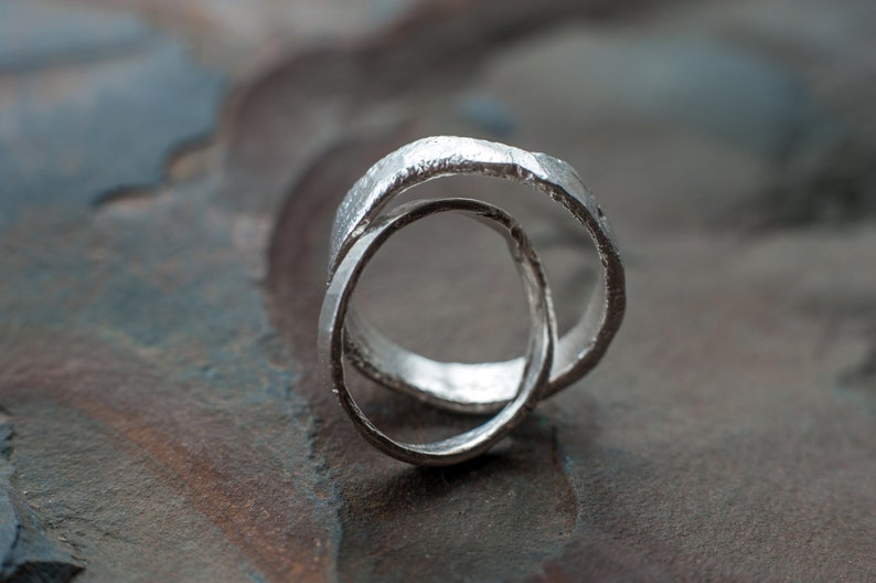 May include: Two silver rings intertwined, creating a unique and modern design. The rings are textured and have a slightly rough finish.