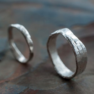 May include: Two silver rings with a textured, hammered finish. The rings are lying on a textured, gray surface.