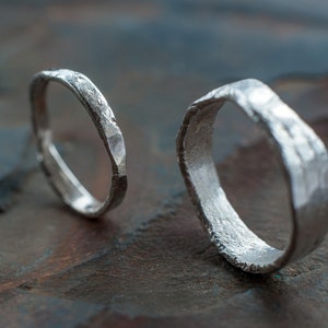 May include: Two silver rings with a hammered texture. The rings are lying on a textured, dark brown surface.