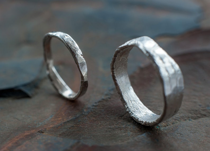 May include: Two silver rings with a hammered texture. The rings are lying on a textured, dark brown surface.
