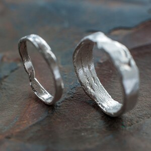 May include: Two silver rings with a textured, hammered finish. The rings are lying on a dark gray, textured surface.