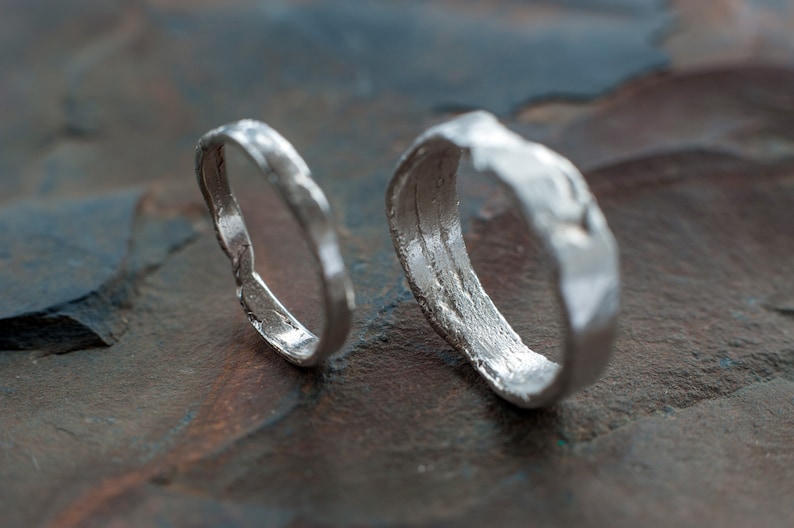 May include: Two silver rings with a textured, hammered finish. The rings are lying on a dark gray, textured surface.