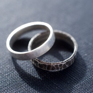 May include: Two silver rings are interlocked. One ring features a smooth, matte, light silver finish with a flat band. The second ring has a dark, oxidized silver finish with a heavily textured, rugged surface that resembles cracked stone or bark.
