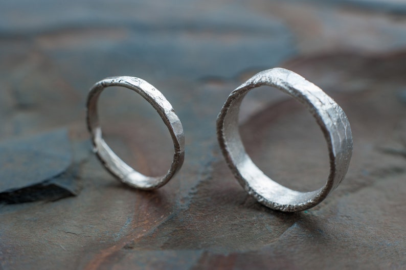 May include: Two silver rings with a hammered texture. The rings are lying on a textured gray surface.