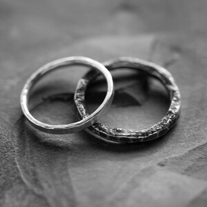 May include: Two silver rings, one with a smooth finish and the other with a textured finish, are intertwined on a gray textured surface.