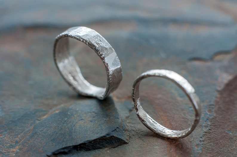 May include: Two silver rings with a textured, hammered finish. The rings are lying on a gray, textured surface.