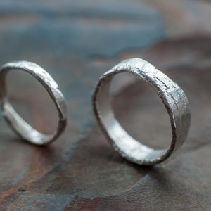 May include: Two silver wedding bands with a hammered texture. The bands are on a textured, dark brown surface.