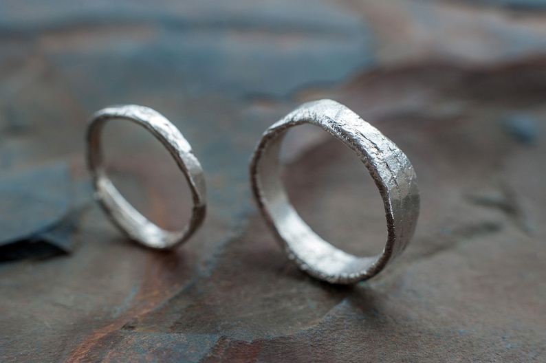 May include: Two silver wedding bands with a hammered texture. The bands are on a textured, dark brown surface.