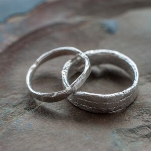 May include: Two silver wedding bands with a textured finish. The bands are intertwined and resting on a gray and brown stone surface.
