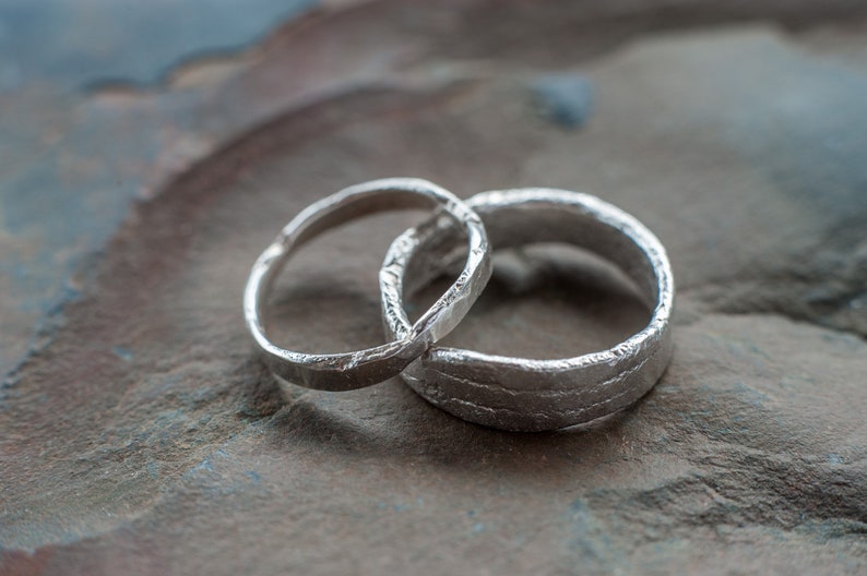 May include: Two silver wedding bands with a textured finish. The bands are intertwined and resting on a gray and brown stone surface.