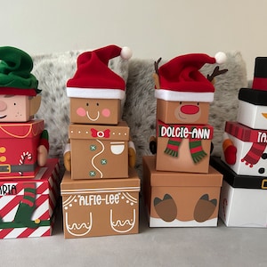 May include: Four stacked Christmas gift boxes with different character themes. The boxes are decorated with festive colours and designs, including red, green, white, and brown. The boxes are labelled with the names "Aria", "Alfie-Lee", "Dolcie-Ann", and "Taylor".