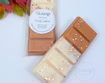 May include: Two wax melt bars with a white and brown color scheme, with gold glitter accents. One bar is in a clear plastic wrapper. The bars have the text "Cosy Cabin" and "Whitcliffe Aromas" printed on them.