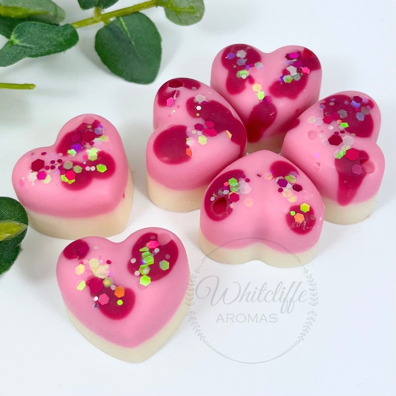 May include: Five heart-shaped wax melts in pink and white with glitter. The wax melts are arranged in a circle on a white surface.
