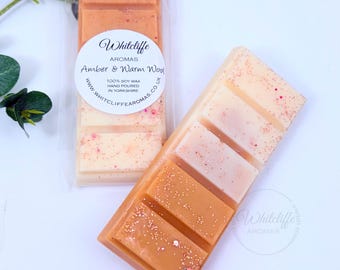 May include: Two wax melt bars with a gradient of colors from light peach to amber. One bar is in a clear plastic wrapper. The bars are labeled "Amber & Warm Wool" from Whitcliffe Aromas, made with 100% soy wax.