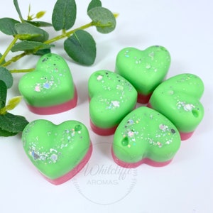 May include: Six heart-shaped wax melts in green and pink with glitter. The wax melts are arranged in a circle on a white surface.