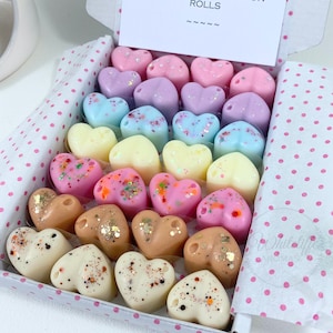 May include: A box filled with heart-shaped wax melts in various colours, including pink, purple, blue, yellow, and brown. Some of the melts have glitter embedded in them. The box is lined with white paper with pink polka dots. The text "Wax Melts Rolls" is printed on the top of the box.