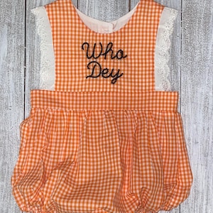 May include: Orange and white gingham baby romper with the words "Who Dey" embroidered in black script. The romper features white lace trim on the shoulders and a gathered, bubble-style bottom. The garment is displayed on a light wood-grain surface.