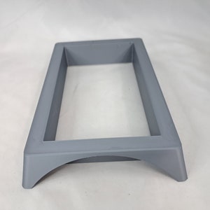 May include: A gray plastic frame with a rectangular opening and a curved base. The frame is designed to hold a small object, such as a phone or a tablet.