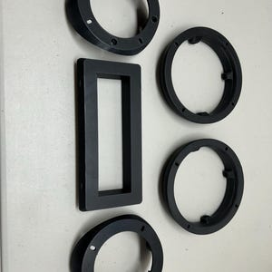 May include: Four black plastic rings with different diameters and a black plastic rectangular frame. The rings have a smooth surface and a raised lip around the edge. The frame has a smooth surface and a raised lip around the edge. The rings and frame are all the same color.