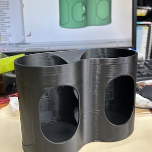 May include: A black 3D-printed cup holder with two circular openings. The holder has a curved design and is made of a textured material. A computer screen in the background displays a green 3D model of the same cup holder.