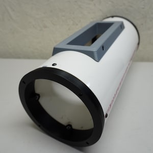 May include: A white cylindrical object with a black circular end and a gray rectangular top. The object has a smooth, glossy finish and appears to be made of plastic or a similar material. The black end has several screw holes.