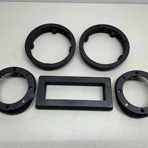 May include: An assortment of black plastic automotive parts, including two circular rings, two smaller circular rings, and a rectangular frame. The parts are arranged on a white surface.