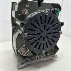 May include: An olive drab electronic device with a black circular vent. The device has an on/off switch, a power button with a green light, and a connector labeled "J2". The device is resting on a gray fabric.