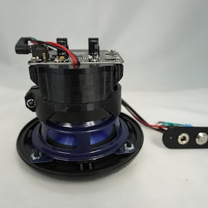 May include: A black speaker with a blue speaker cone and a circuit board attached to the top. The speaker has a black plastic housing and a black wire coming out of the back. There is a separate black plastic piece with two silver jacks.