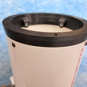 May include: Close-up of a white telescope tube with a black ring around the top. The ring has four small black screws and a small hole in the center.