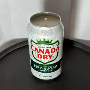 May include: A candle in a Canada Dry Zero Sugar Ginger Ale can. The can is white with green and red accents, and the text "CANADA DRY" is prominently displayed. The candle has a silver rim and a white wick.