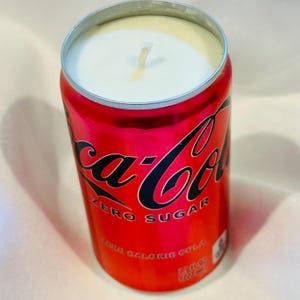 May include: A red Coca-Cola Zero Sugar can candle with a white wax interior and a wick. The can features the Coca-Cola logo in black and white, with the words "ZERO SUGAR" and "ZERO CALORIE COLA".