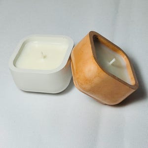 May include: Two small, square candles with white wax and wicks. One candle is in a white, square container, and the other is in a tan, geometric-shaped container. The candles are on a white surface.