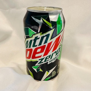May include: A Mountain Dew Zero Sugar can with a black background and green and white geometric shapes. The words "Mtn Dew" are in white and red, with "Zero Sugar" in green. The can is on a white surface.