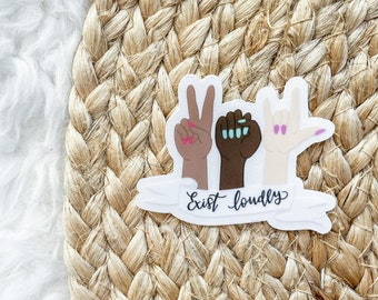 Puede incluir: Una pegatina con tres manos de diferentes tonos de piel, cada una mostrando un gesto: paz, puño y rock on. El texto "Exist loudly" está escrito en cursiva debajo. La pegatina está sobre un fondo tejido.