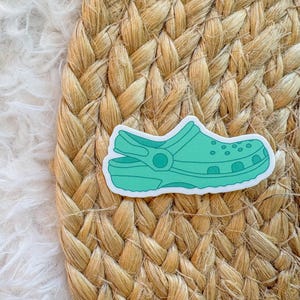 Puede incluir: Una pegatina turquesa con forma de zueco, con un borde blanco. La pegatina está colocada sobre una superficie tejida de color marrón claro. El fondo incluye una textura blanca y esponjosa.