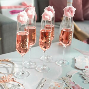 Puede incluir: Algod&oacute;n de az&uacute;car rosa se sienta encima de copas de champ&aacute;n llenas de champ&aacute;n rosado. Cada copa tiene una pegatina dorada que dice "Bride Tribe".
