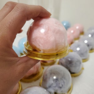 Puede incluir: Primer plano de una bola de algod&oacute;n de az&uacute;car rosa en una c&uacute;pula de pl&aacute;stico transparente con un borde dorado. La c&uacute;pula est&aacute; sobre una superficie blanca. Hay otras bolas de algod&oacute;n de az&uacute;car en el fondo, algunas son rosas y otras son moradas.