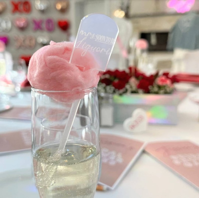 Puede incluir: Un palo de pl&aacute;stico transparente con una bola de algod&oacute;n de az&uacute;car rosa en la parte superior. El palo tiene un cartel blanco pegado que dice "Mrs. Soon to be Ligouri".