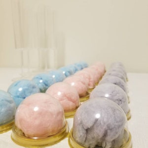 Puede incluir: Una fila de recipientes de pl&aacute;stico transparente llenos de algod&oacute;n de az&uacute;car de color pastel. Los recipientes tienen tapas doradas y est&aacute;n dispuestos en l&iacute;nea sobre una superficie blanca.