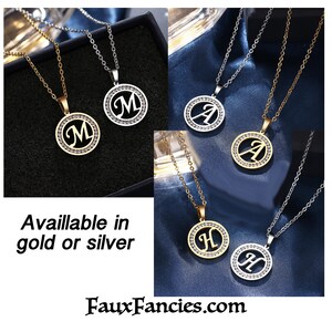 May include: Gold and silver-toned circular pendant necklaces featuring the letters M, A, and H. Each pendant has a central cutout letter surrounded by a ring of small clear crystals on a matching metal chain. Text reads: Available in gold or silver.