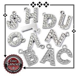 May include: Silver-toned alphabet charms, each encrusted with clear crystals. The letters include S, H, D, U, O, A, N, B, A, and C. The charms are displayed on a white background with a black border. The logo "Faux Fancies" is visible.
