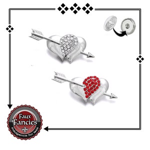 May include: Two silver heart-shaped brooches with arrows through them. One brooch has clear rhinestones and the other has red rhinestones. The brooches have a pin back with a magnetic closure.