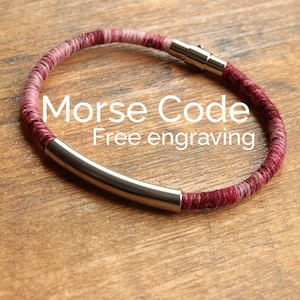 May include: A red and pink braided bracelet with a silver metal bar and a clasp. The bracelet has the text "Morse Code Free engraving" on it.