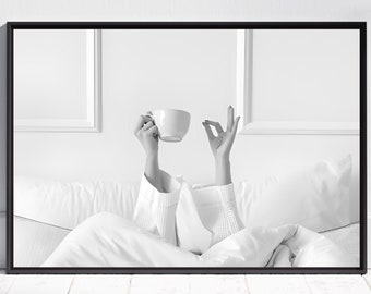 May include: A black and white photograph of a person in bed holding a white mug. The person is wearing a white sweater and has their hands raised in a relaxed pose. The bed is made with white sheets and pillows.