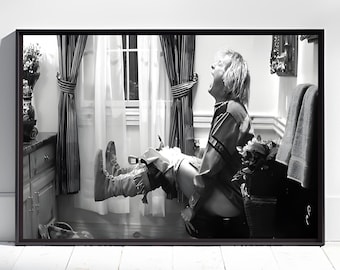 May include: Black and white framed photograph of a person in a bathroom, seated on a toilet with their legs extended. The person is wearing a shirt and boots, with an open mouth. The bathroom has a window with curtains.