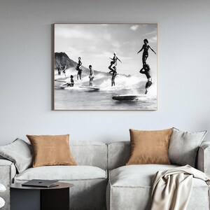 May include: Black and white photograph of a group of people surfing on a beach. The people are standing on surfboards and are in the process of riding waves. The photo is taken from a distance and shows the people in the water with the beach in the background.